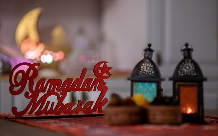 Dekoratives Arrangement mit zwei orientalischen Laternen und einem Schriftzug aus Holz, der „Ramadan Mubarak“ zeigt. Im Hintergrund leuchten unscharfe Lichter in warmen Farben.