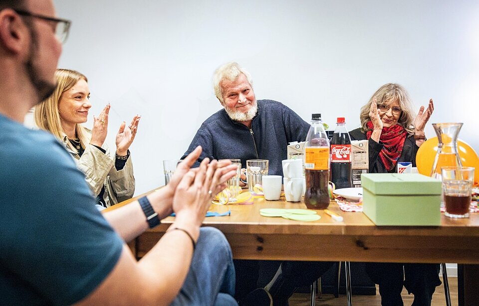 Eine Gruppe von Menschen sitzt um einen Tisch und applaudiert. Im Vordergrund klatscht eine Person in die Hände. In der Mitte sitzt ein älterer Mann mit weißem Bart, der lächelt und ebenfalls applaudiert. Rechts klatscht eine Frau mit Brille und rotem Schal. Auf dem Tisch sind verschiedene Getränke, darunter Coca-Cola und Saft, zu sehen. Außerdem befindet sich auf dem Tisch ein grüner Pappkarton mit Schlitz im Deckel.