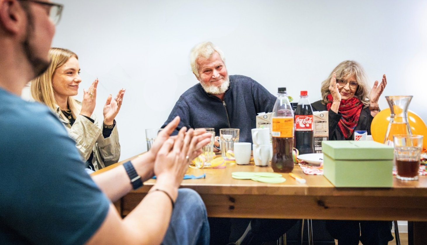 Eine Gruppe von Menschen sitzt um einen Tisch und applaudiert. Im Vordergrund klatscht eine Person in die Hände. In der Mitte sitzt ein älterer Mann mit weißem Bart, der lächelt und ebenfalls applaudiert. Rechts klatscht eine Frau mit Brille und rotem Schal. Auf dem Tisch sind verschiedene Getränke, darunter Coca-Cola und Saft, zu sehen. Außerdem befindet sich auf dem Tisch ein grüner Pappkarton mit Schlitz im Deckel.