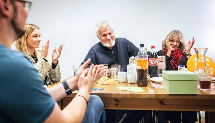 Eine Gruppe von Menschen sitzt um einen Tisch und applaudiert. Im Vordergrund klatscht eine Person in die Hände. In der Mitte sitzt ein älterer Mann mit weißem Bart, der lächelt und ebenfalls applaudiert. Rechts klatscht eine Frau mit Brille und rotem Schal. Auf dem Tisch sind verschiedene Getränke, darunter Coca-Cola und Saft, zu sehen. Außerdem befindet sich auf dem Tisch ein grüner Pappkarton mit Schlitz im Deckel.