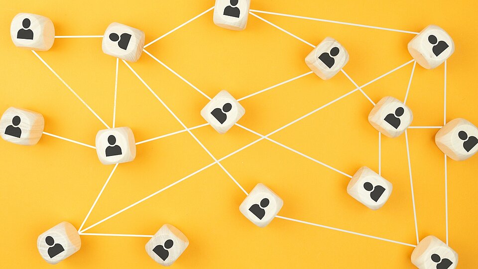 Holzwürfel mit Personen-Symbolen sind über weiße Linien auf gelbem Hintergrund miteinander verbunden und visualisieren ein Netzwerk aus Beziehungen und Kooperationen.
