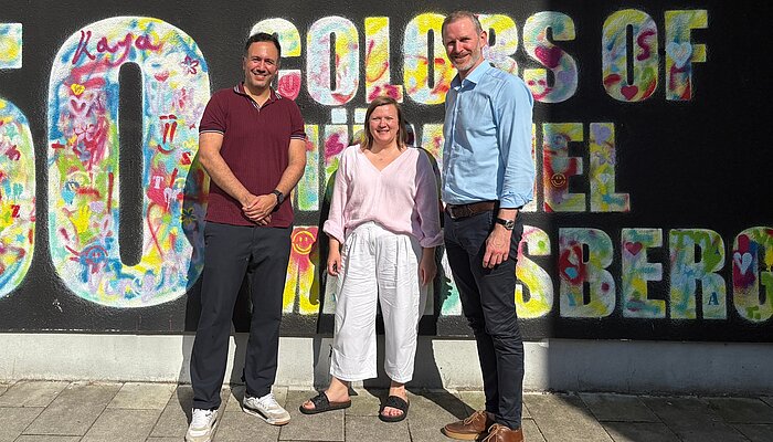 Baris Önes, Oksana Umland und Simon Steinwachs stehen vor einer bunt gestalteten Street-Art-Wand in Mümmelmannsberg.