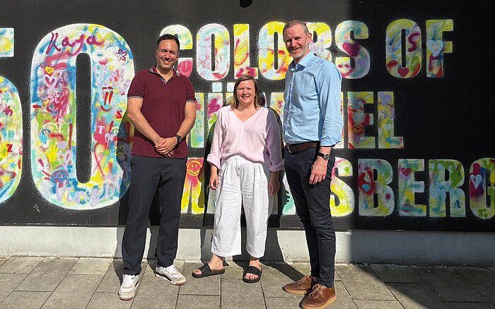 Baris Önes, Oksana Umland und Simon Steinwachs stehen vor einer bunt gestalteten Street-Art-Wand in Mümmelmannsberg.