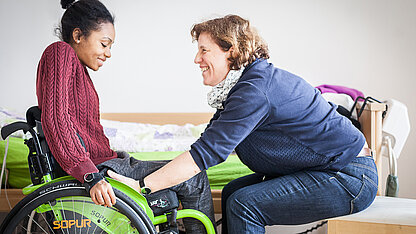 Eine junge Person im Rollstuhl wird von einer Assistenzkraft freundlich unterstützt. Die Assistenzkraft geht behutsam vor und hält die Hände an die der jungen Person, um sie beim Aufstehen oder Umsetzen zu unterstützen. Beide lächeln einander an.