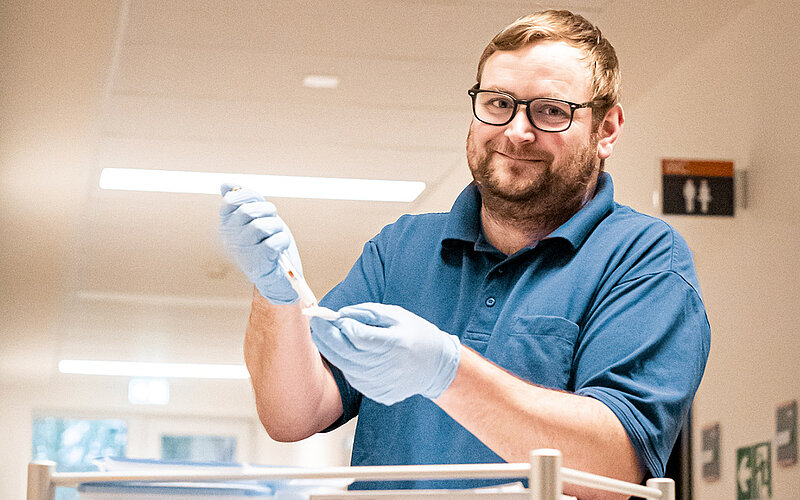 Ein Pfleger mit Brille und blauer Kleidung trägt Handschuhe und hält eine medizinische Spritze. Er lächelt freundlich und steht in einem hellen Raum.