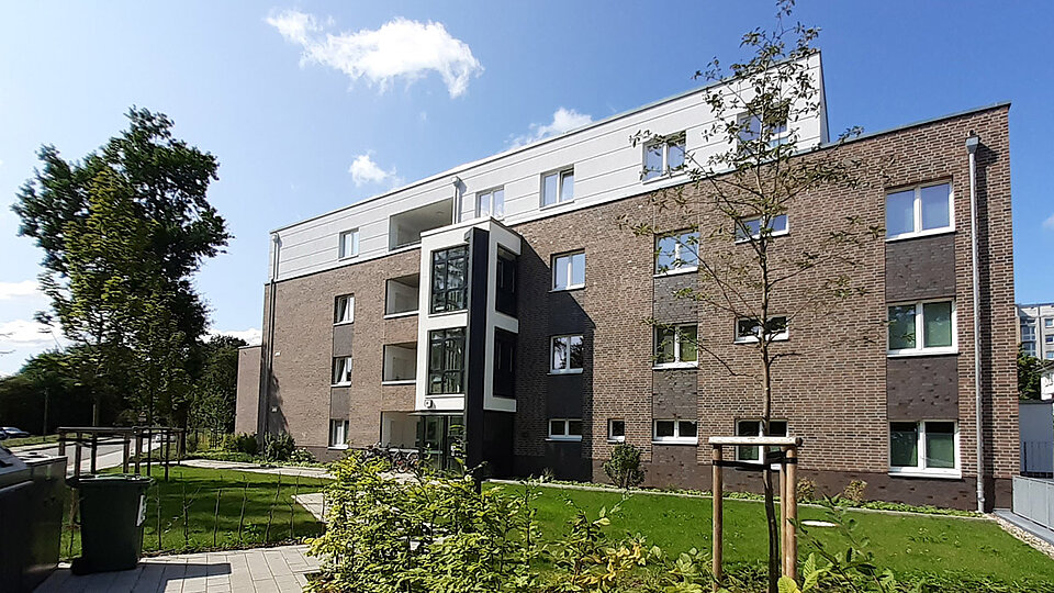Ein modernes Wohngebäude mit dunkler Klinkerfassade und weißen Akzenten steht auf einem begrünten Grundstück mit jungen Bäumen. Der Zugang ist barrierefrei.