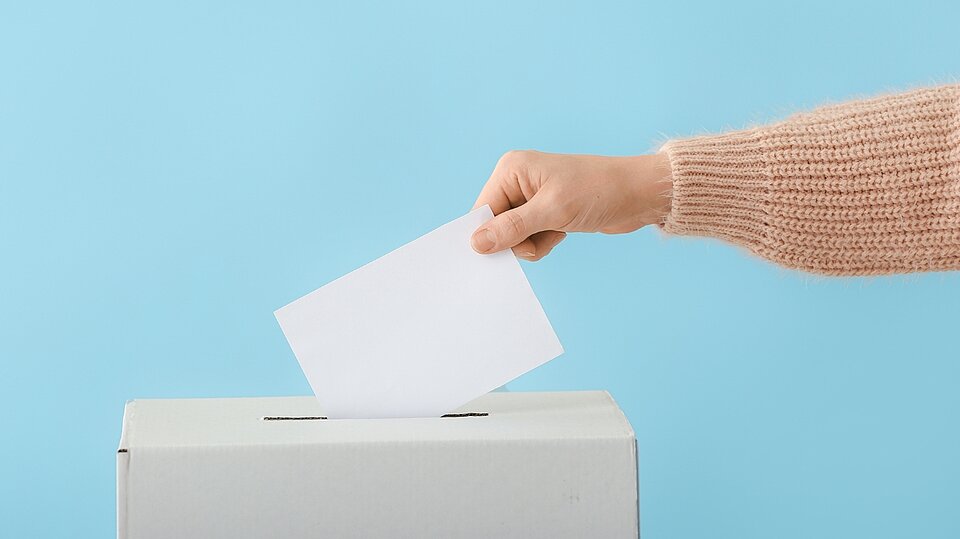 Eine Person steckt einen Stimmzettel in eine Wahlurne.