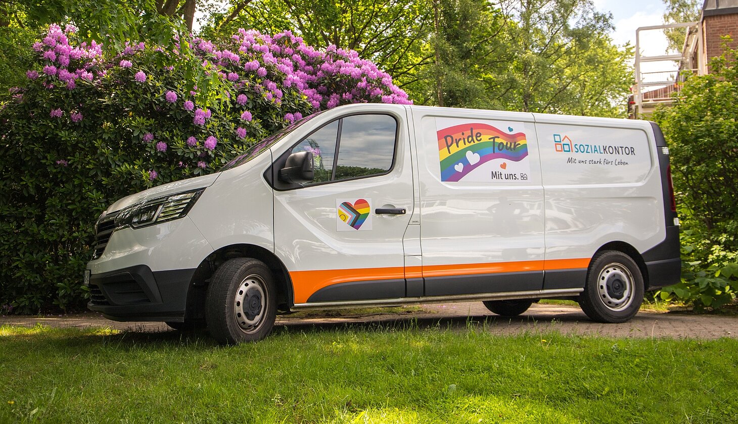 Der Pride-Transporter des Sozialkontors steht auf einem grünen Grundstück vor blühenden Rhododendren. Auf der Fahrzeugseite sind das Logo der Pride Tour und das Sozialkontor-Logo zu sehen.