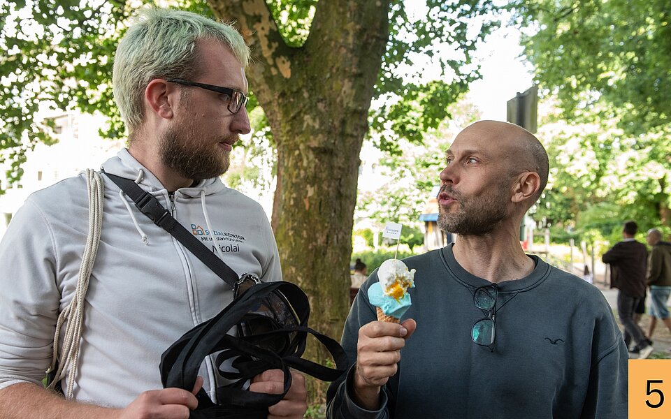 Ein Mitarbeiter des Sozialkontors spricht unter einem Baum mit einem Besucher des Stadtteilfests, der eine Eiswaffel in der Hand hält.