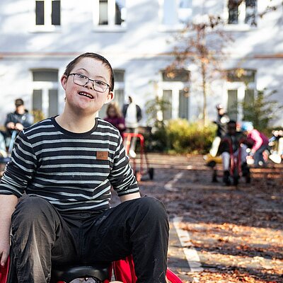 Ein Förderschüler im Rollstuhl blickt in die Kamera, hinter ihm ein hellblaues Haus und verschwommen weitere Kinder mit Behinderung.