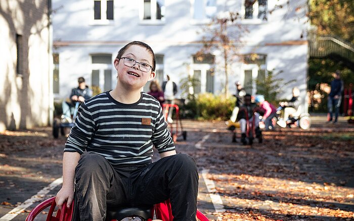 Ein Schüler einer Förderschule im Rollstuhl vor einem hellblauen Haus, im Hintergrund unscharf weitere Kinder mit Behinderung