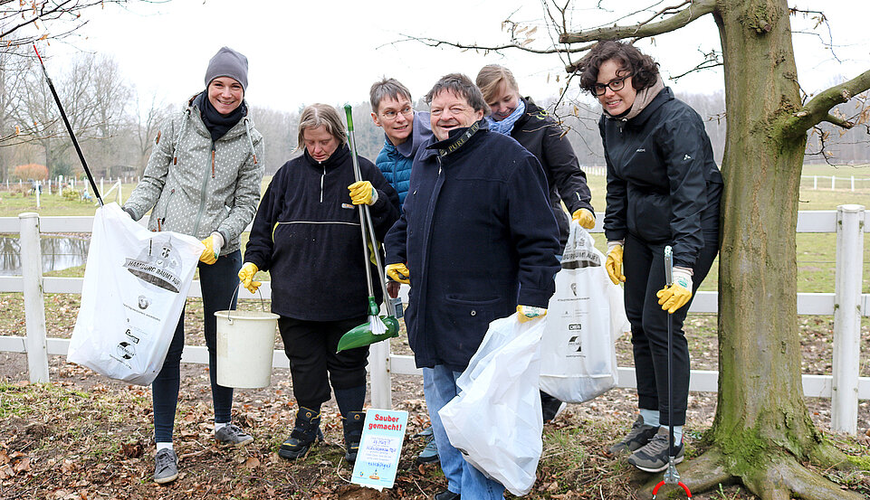 Im Freien. Sechs Personen beim Müllaufsammeln, ausgestattet mit Mülltüten, Eimern und Greifzangen.