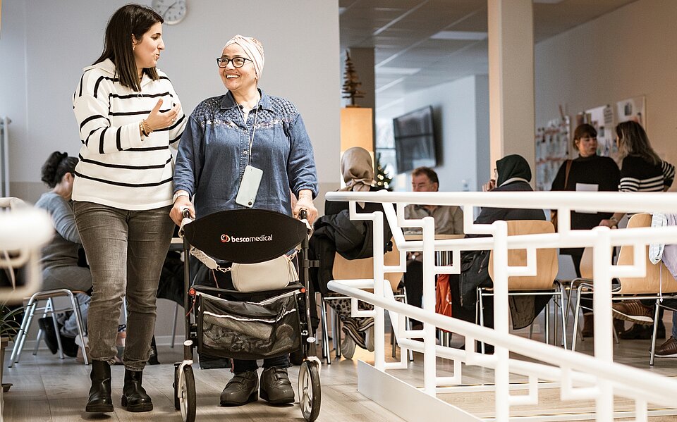 Treffpunkte im Sozialkontor: Zwei Frauen, eine davon mit Rollator, unterhalten sich. Im Hintergrund sitzen weitere Menschen an Tischen.