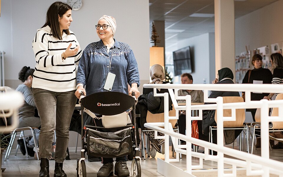 Treffpunkte im Sozialkontor: Zwei Frauen, eine davon mit Rollator, unterhalten sich. Im Hintergrund sitzen weitere Menschen an Tischen.