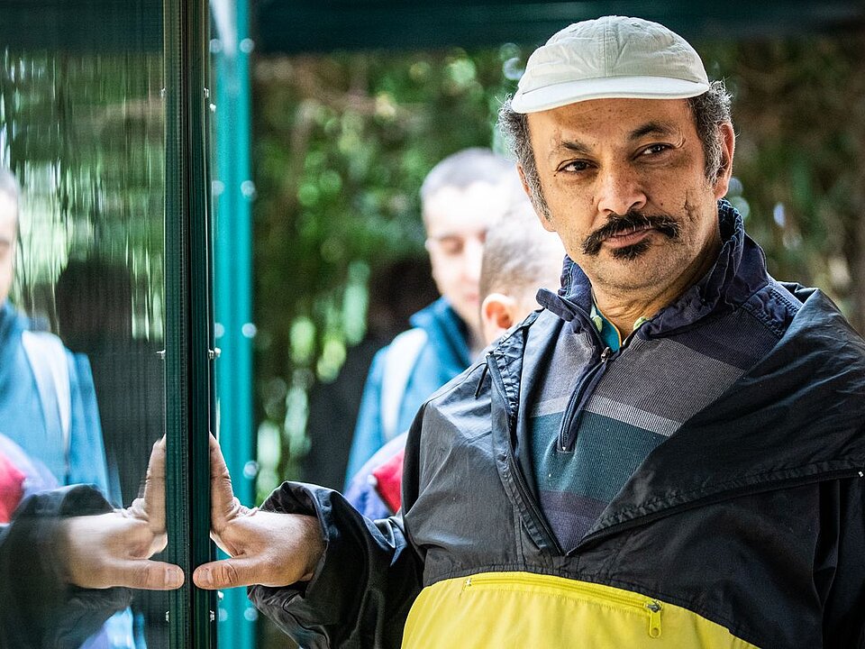 Ein Mann mit Kappe steht vor einer Glasscheibe und macht eine einladende Geste. Seine Spiegelung ist im Glas sichtbar. Im Hintergrund sind weitere Menschen zu sehen. 