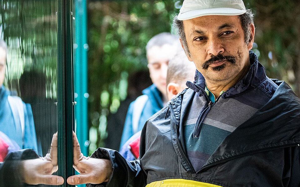 Ein Mann mit Kappe steht vor einer Glasscheibe und macht eine einladende Geste. Seine Spiegelung ist im Glas sichtbar. Im Hintergrund sind weitere Menschen zu sehen. 