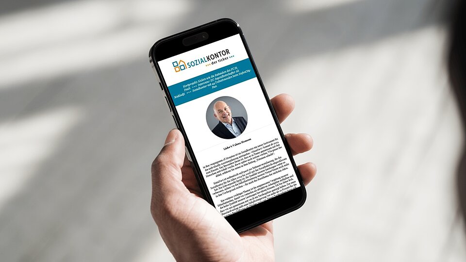 Eine Person hält ein Smartphone in der Hand. Auf dem Bildschirm ist ein geöffneter Newsletter des Sozialkontors zu sehen. Darunter ist ein Bild und ein Textabschnitt sichtbar. Der Hintergrund ist unscharf und zeigt helle Licht- und Schattenspiele.