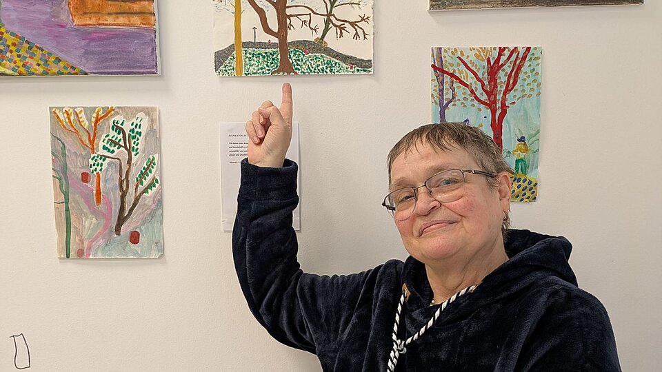 Frau zeigt auf ihre selbstgemalten Bilder an der Wand