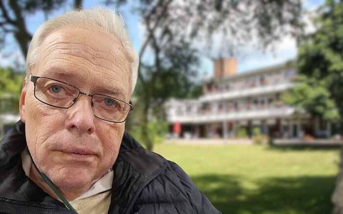 Roland Tessner im Vordergrund, mit Brille und freundlichem Blick. Im Hintergrund das Senator-Neumann-Haus, umgeben von einer grünen Wiese und Bäumen.