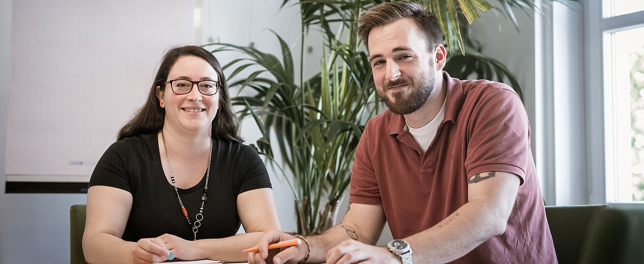 Zwei Personen sitzen nebeneinander an einem Tisch und blicken freundlich in die Kamera. Auf dem Tisch liegen Schreibutensilien. Im Hintergrund stehen große Pflanzen.