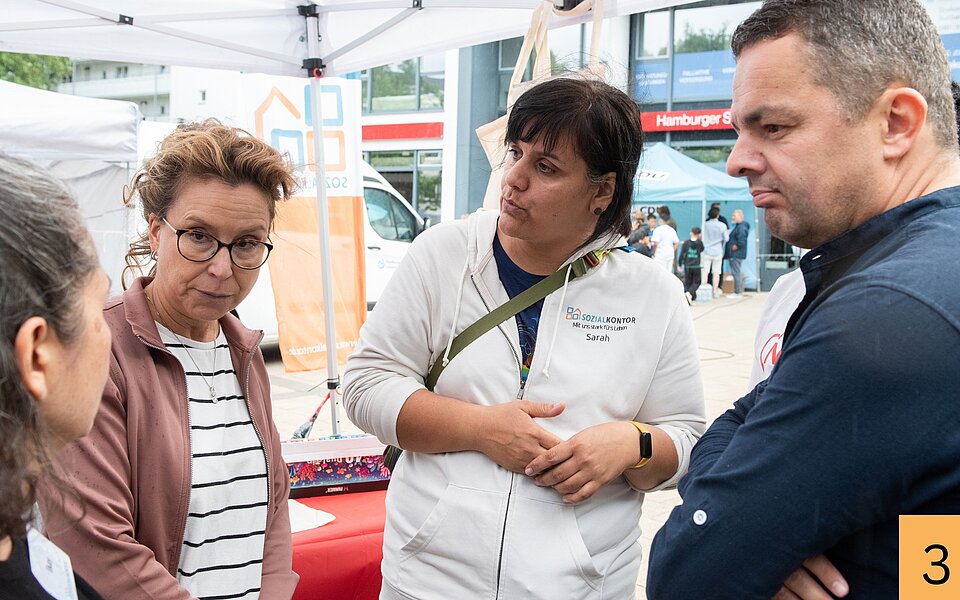 Carola Veit und Ralf Neubauer stehen beim Stadtteilfest Rothenburgsort mit Mitarbeiterinnen des Sozialkontors unter einem Pavillon und sind miteinander im Gespräch.
