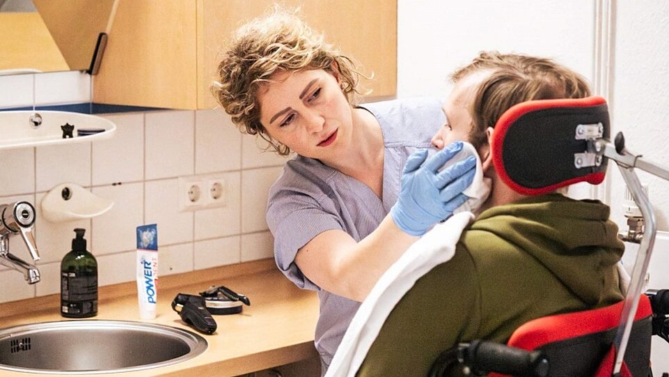 Eine Person in Pflegekleidung reinigt mit einem Tuch das Gesicht einer Person, die in einem Rollstuhl sitzt. Die Szene spielt in einem Badezimmer mit Waschbecken und Pflegeutensilien auf der Ablage.