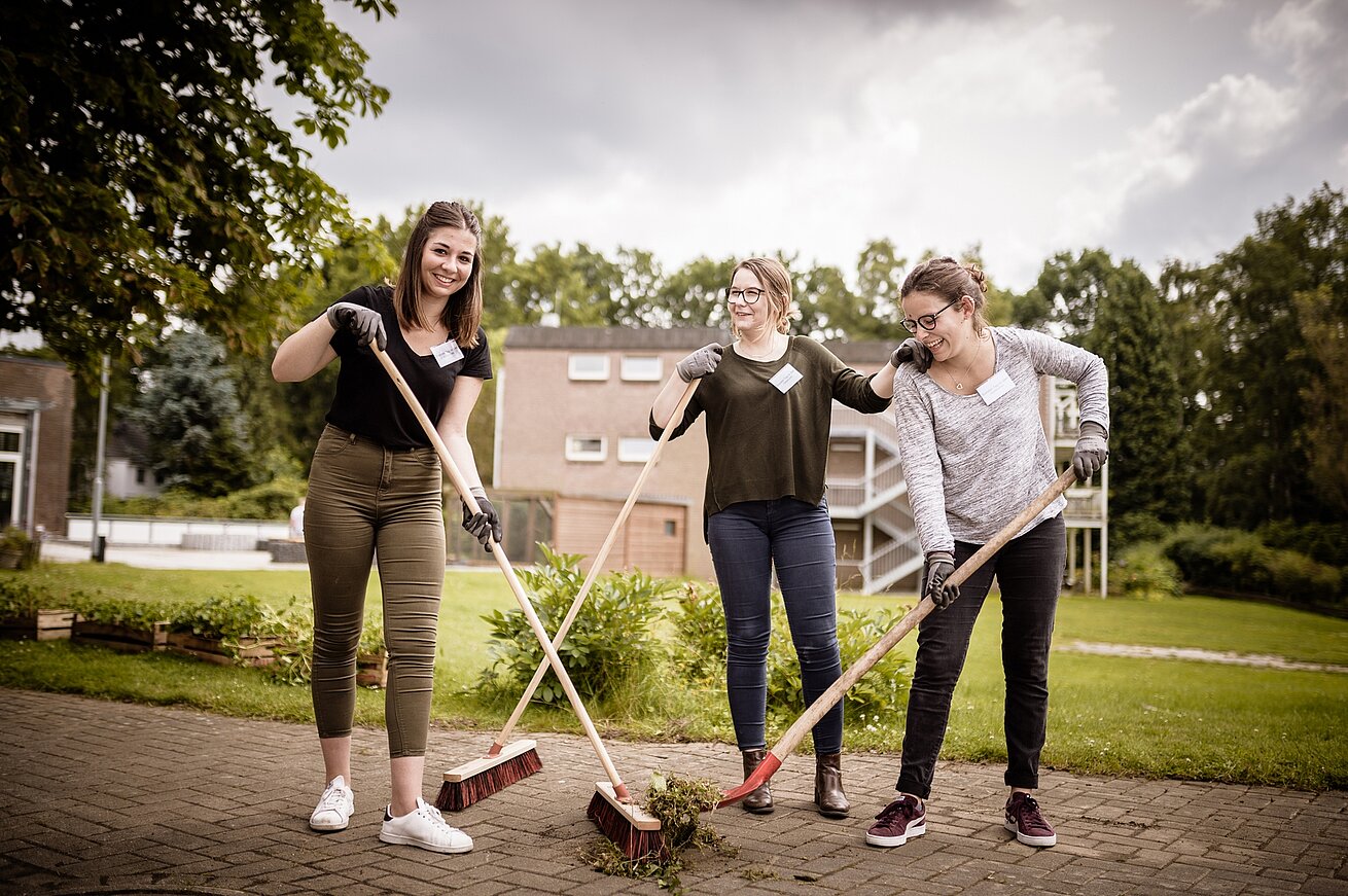 Im Freien. Drei junge Frauen mit Besen und Schaufel bei der Gartenarbeit.