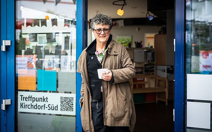 Frau steht mit einer Tasse in der Hand am Eingang eines Gebäudes, an der Tür sind ein QR-Code und ein Schild mit der Aufschrift „Treffpunkt Kirchdorf-Süd“ zu sehen.