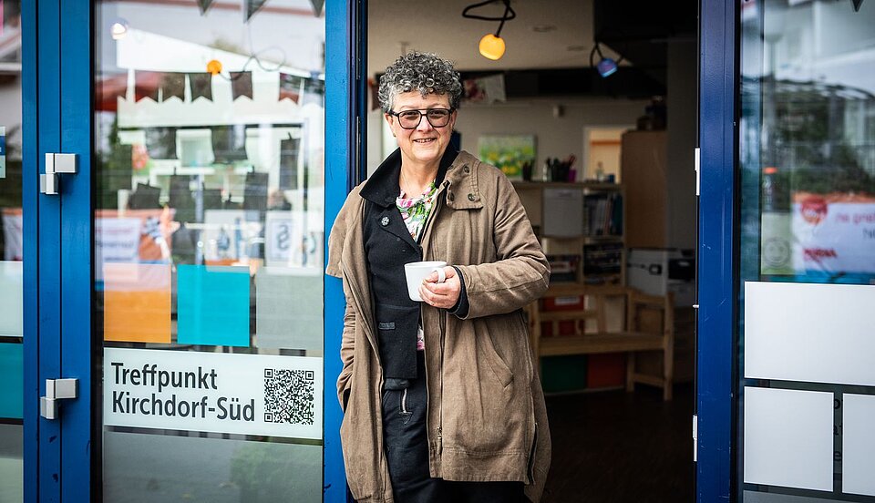 Frau steht mit einer Tasse in der Hand am Eingang eines Gebäudes, an der Tür sind ein QR-Code und ein Schild mit der Aufschrift „Treffpunkt Kirchdorf-Süd“ zu sehen.
