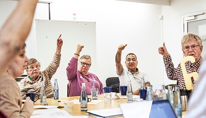 Mehrere Menschen sitzen an einem Tisch in einem Workshop und stimmen mit erhobenem Daumen über eine Frage ab.