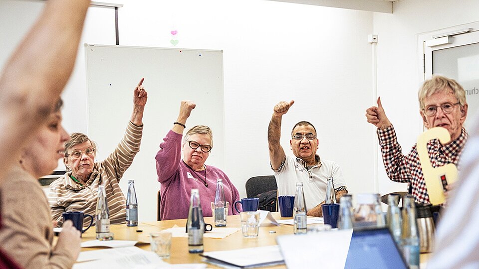 Mehrere Menschen sitzen an einem Tisch in einem Workshop und stimmen mit erhobenem Daumen über eine Frage ab.
