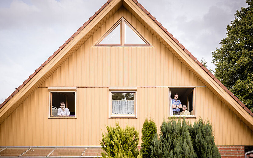 Ein gelbes Wohnhaus mit Satteldach und drei geöffneten Fenstern im oberen Stockwerk. Aus zwei Fenstern schauen insgesamt drei Personen lächelnd hinaus. Im Vordergrund wachsen grüne Sträucher. 