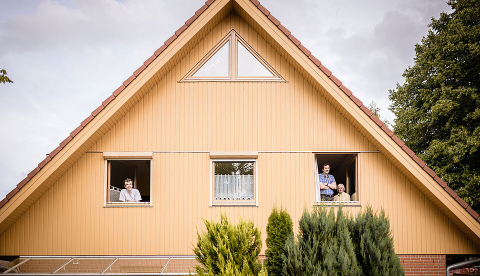 Ein gelbes Wohnhaus mit Satteldach und drei geöffneten Fenstern im oberen Stockwerk. Aus zwei Fenstern schauen insgesamt drei Personen lächelnd hinaus. Im Vordergrund wachsen grüne Sträucher.