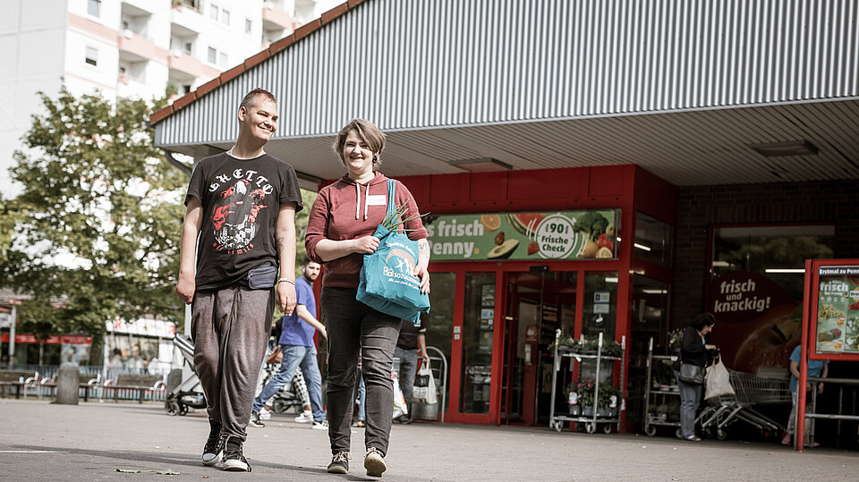 Zwei Menschen kommen lachend aus einem Supermarkt und tragen Einkaufstaschen. Im Hintergrund sind weitere Personen und Einkaufswagen zu sehen.