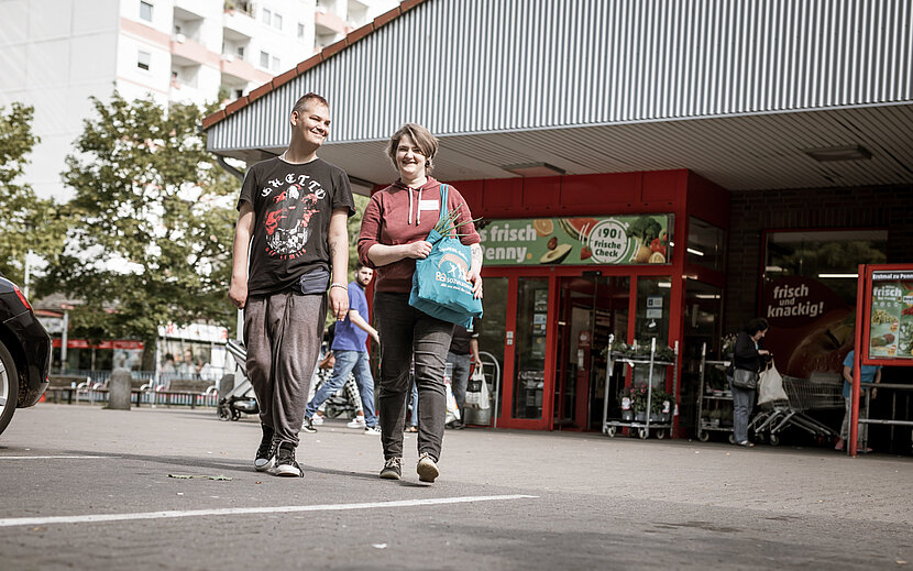 Zwei Menschen kommen lachend aus einem Supermarkt und tragen Einkaufstaschen. Im Hintergrund sind weitere Personen und Einkaufswagen zu sehen.