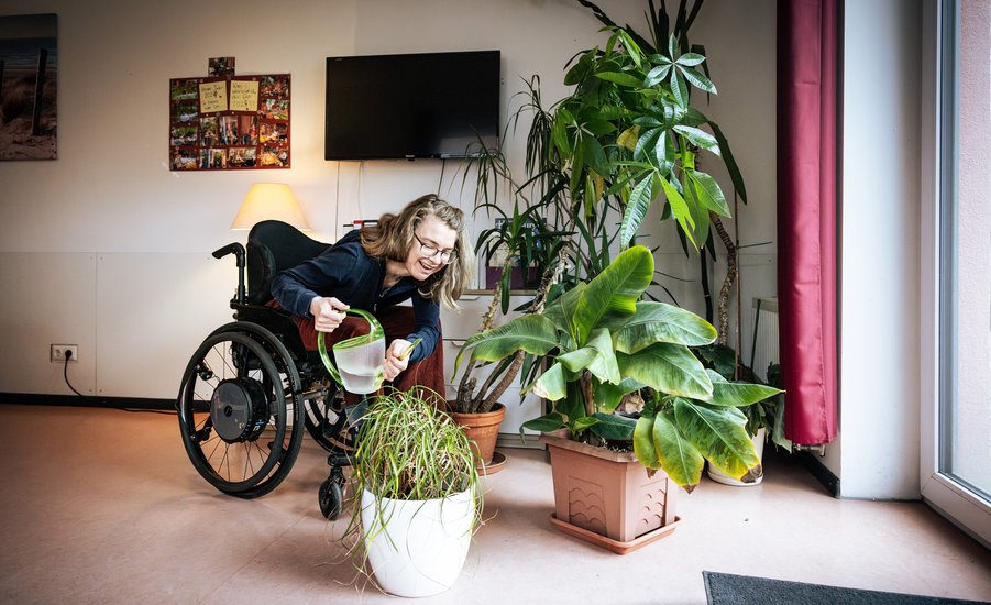 Eine Frau im Rollstuhl gießt mit einer grünen Gießkanne Zimmerpflanzen in einem hellen Raum. Sie beugt sich mit einem Lächeln nach vorn zu einem großen, weißen Blumentopf. Um sie herum stehen weitere grüne Pflanzen. Im Hintergrund sind eine Stehlampe, ein Fernseher, eine Fotowand und ein Fenster mit rotem Vorhang zu sehen.
