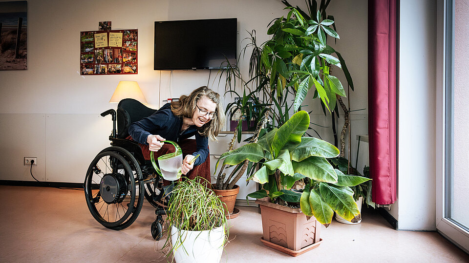 Eine Frau im Rollstuhl gießt mit einer grünen Gießkanne Zimmerpflanzen in einem hellen Raum. Sie beugt sich mit einem Lächeln nach vorn zu einem großen, weißen Blumentopf. Um sie herum stehen weitere grüne Pflanzen. Im Hintergrund sind eine Stehlampe, ein Fernseher, eine Fotowand und ein Fenster mit rotem Vorhang zu sehen.