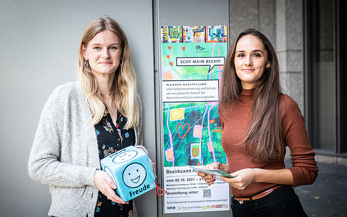 Zwei Frauen stehen nebeneinander, die eine hält einen Würfel mit Emotionen und die andere hält Flyer für die