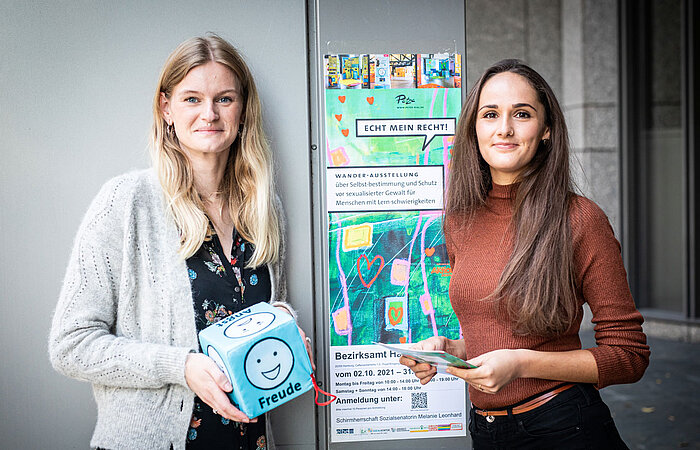 Zwei Frauen stehen nebeneinander, die eine hält einen Würfel mit Emotionen und die andere hält Flyer für die 