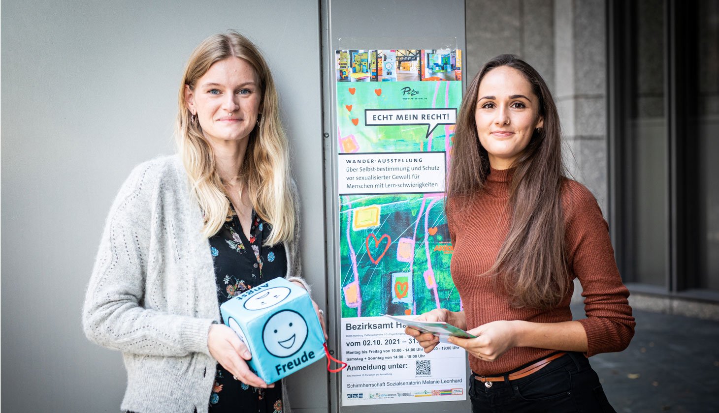 Zwei Frauen stehen nebeneinander, die eine hält einen Würfel mit Emotionen und die andere hält Flyer für die 