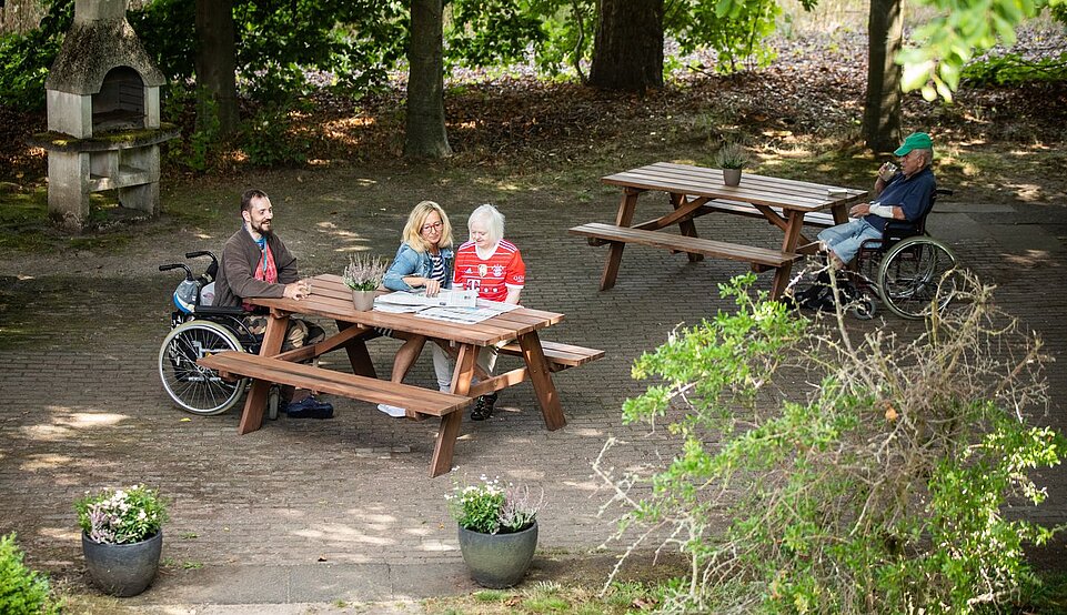 Drei Menschen sitzen an einem Picknick-Tisch und ein weiterer Mensch sitzt hinter ihnen an einem anderen Tisch