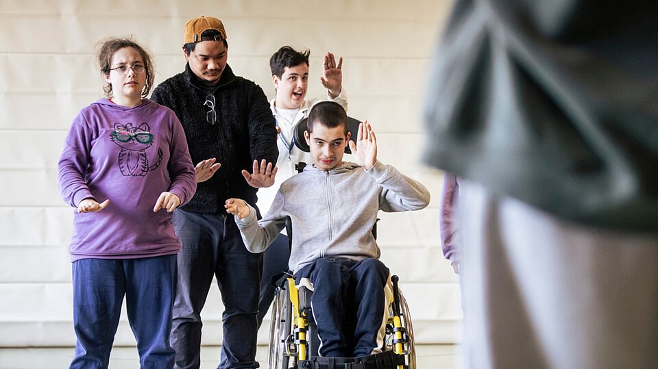 : Mehrere Schüler*innen der schulischen Ganztagsbetreuung des Sozialkontors bewegen sich gemeinsam bei einem Hip-Hop-Workshop in einer Turnhalle. Ein Jugendlicher im Rollstuhl und weitere Kinder folgen aufmerksam einer Tanzanleitung.