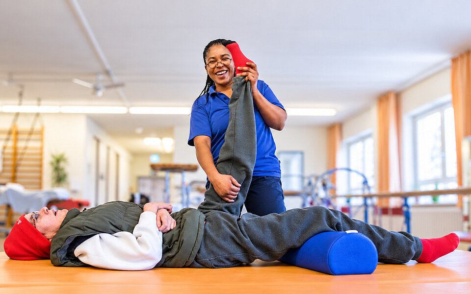 Therapiezentrum Sozialkontor: Eine lächelnde Therapeutin unterstützt eine liegende Person bei einer Beinübung auf einer Therapieliege.