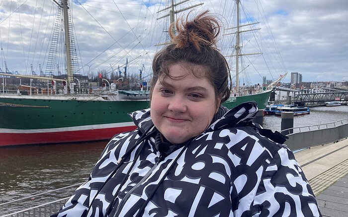 Eine lächelnde Frau sitzt im Rollstuhl am Hamburger Hafen, im Hintergrund ist ein Schiff zu sehen.