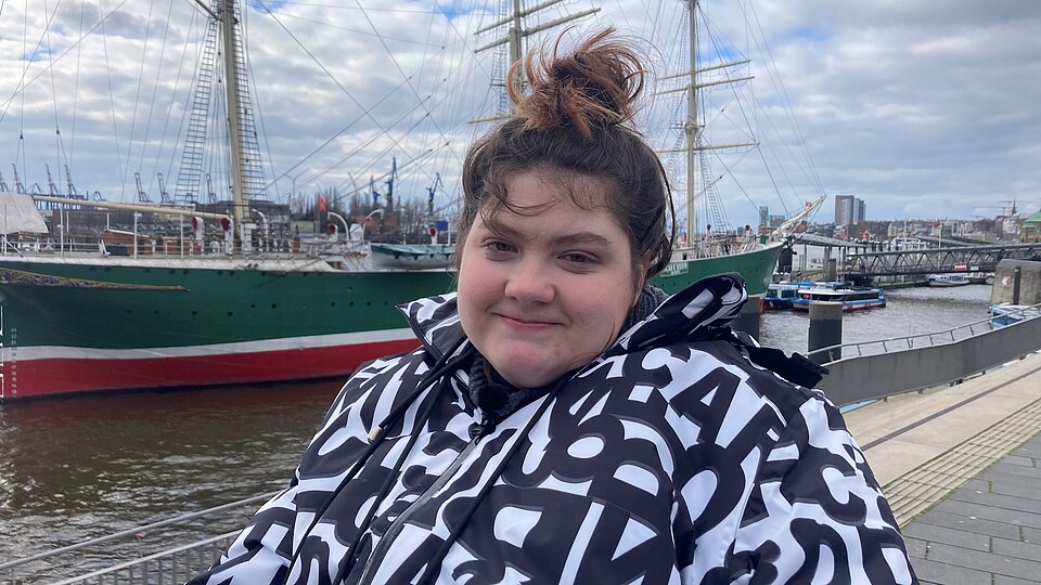 Eine lächelnde Frau sitzt im Rollstuhl am Hamburger Hafen, im Hintergrund ist ein Schiff zu sehen.