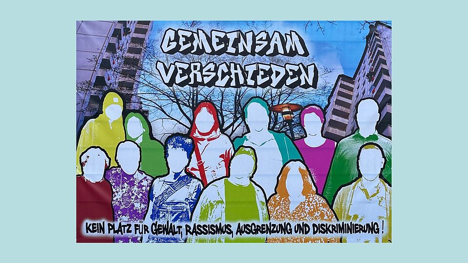 Graffiti-ähnliches Wandbild mit vielen bunten, stilisierten Menschen ohne erkennbare Gesichtszüge. Oben steht „Gemeinsam verschieden“, unten: „Kein Platz für Gewalt, Rassismus, Ausgrenzung und Diskriminierung!“. Im Hintergrund sind Wohngebäude zu sehen.