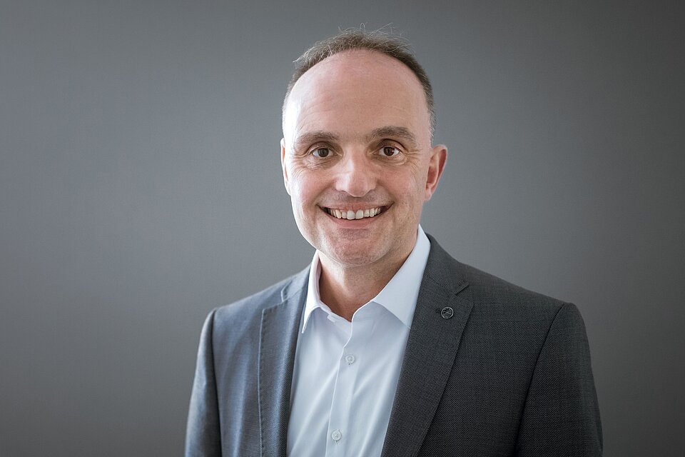 Ein lächelnder Mann in einem Anzug steht vor einer grauen Wand. Der Mann ist Michael Wild, Leiter Strategie & Organisationsentwicklung beim Sozialkontor.