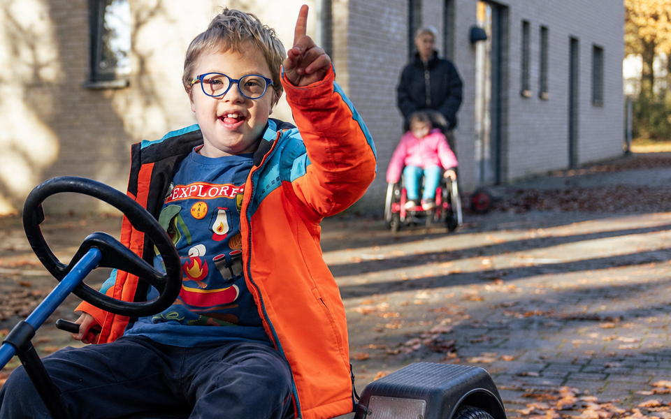 Schulischer Ganztag im Sozialkontor: Ein lächelndes Kind mit Brille und orangefarbener Jacke sitzt auf einem Kettcar und zeigt nach oben. Im Hintergrund sind ein weiteres Kind im Rollstuhl und eine erwachsene Person zu sehen.