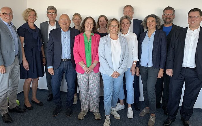 Eine Gruppe von 13 Erwachsenen in Berufskleidung posiert gemeinsam für ein Foto in einer Büroumgebung.