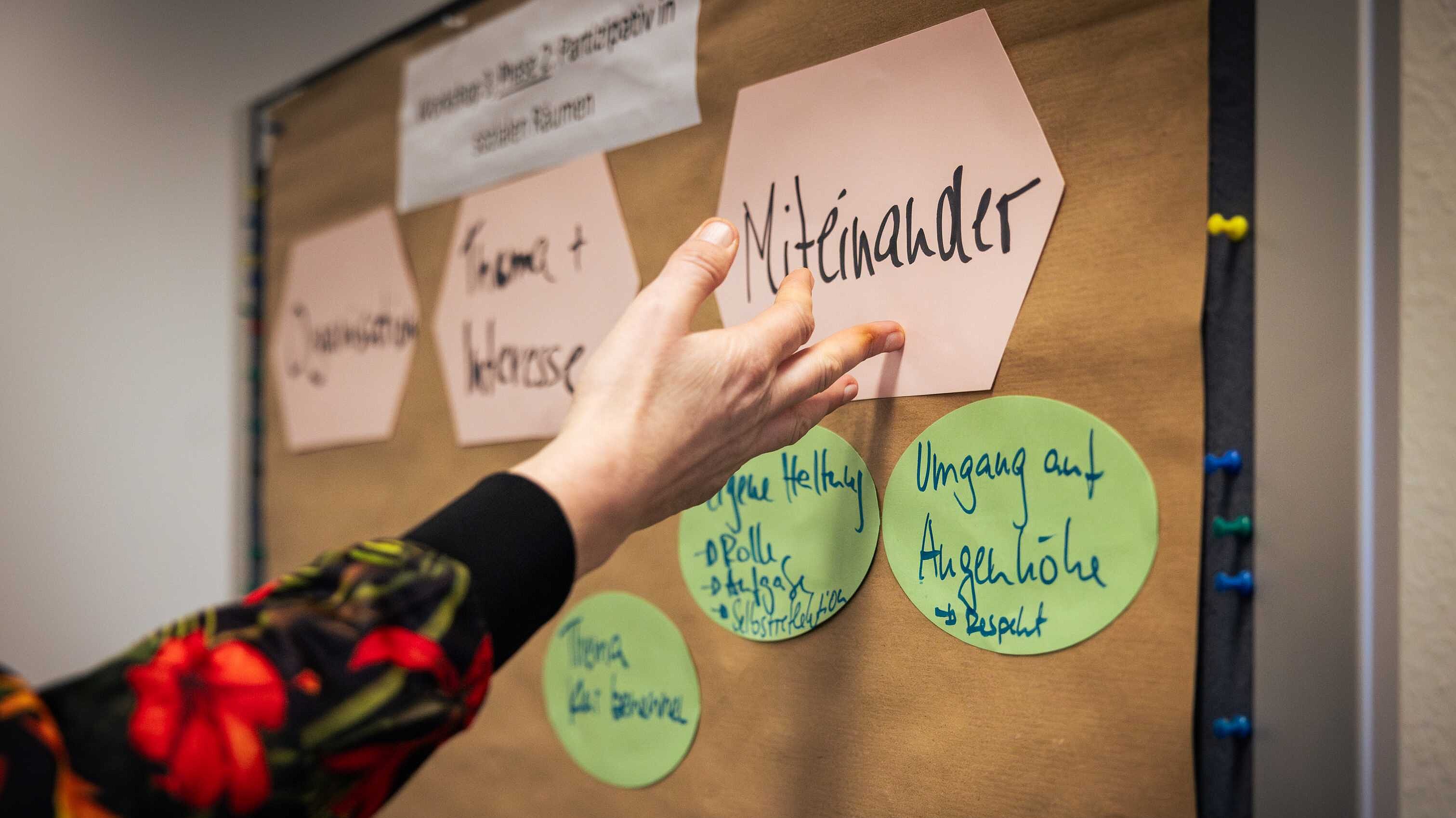 Eine Person zeigt auf eine eine rosa, sechseckige Karte mit dem Wort „Miteinander“ auf einer Pinnwand. Links daneben hängen zwei weitere rosa Sechsecke mit den Worten „Organisation“ und „Thema + Interesse“. Mittig über den rosa Karten steht auf einer weißen Karte die Überschrift „Workshop 3 Phase 2: Partizipation in sozialen Räumen“.  Unter der Karte mit dem Wort „Miteinander“ sind zwei grüne, runde Karten zu sehen. Die erste grüne Karte trägt die Aufschrift: „Eigene Haltung – Rolle – Auftrag – Selbstreflexion“. Die zweite: „Umgang auf Augenhöhe – Respekt“.